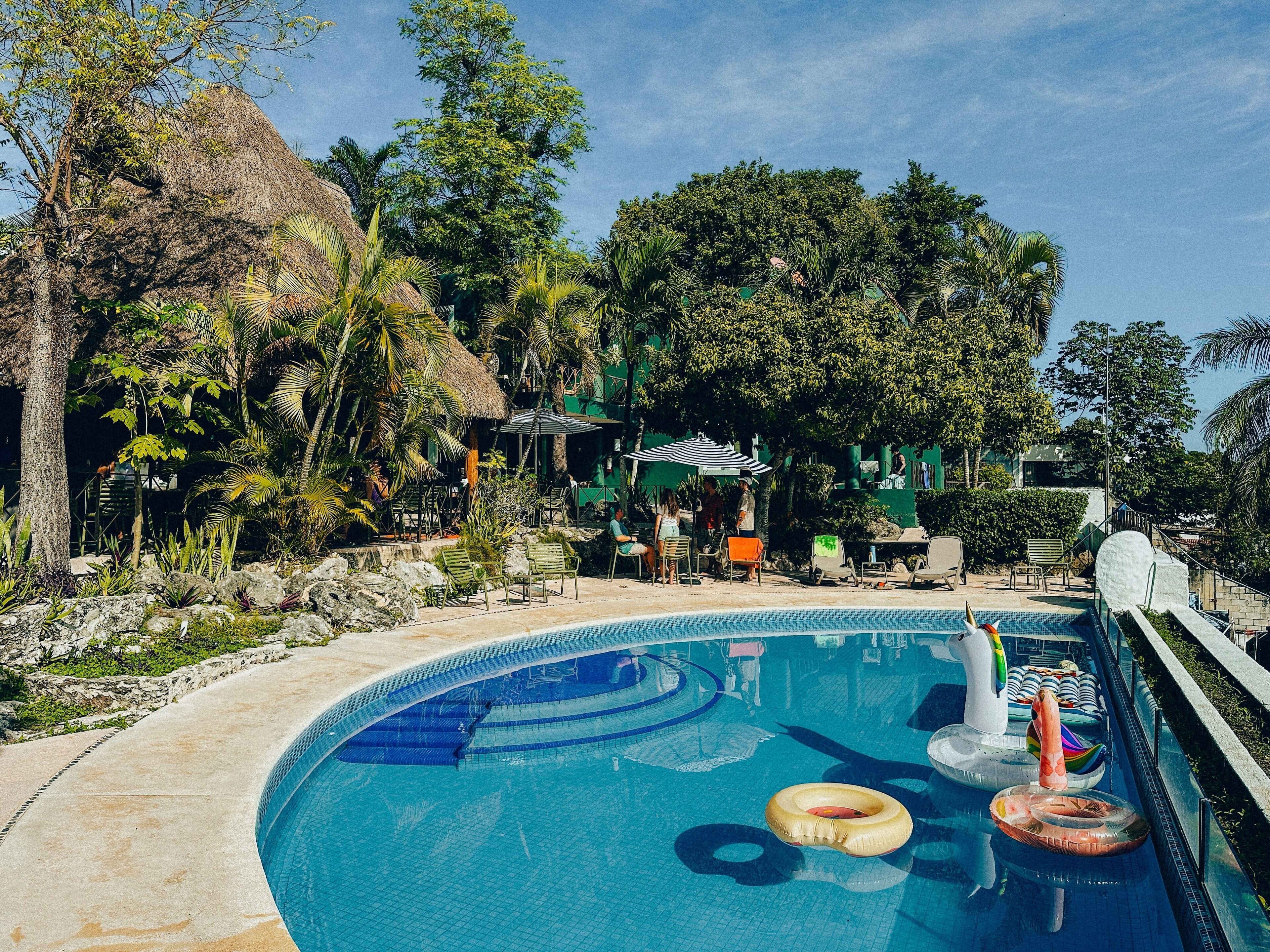 A pool with lounge chairs and inflatable rings, surrounded by tropical vegetation and a building with a thatched roof.
