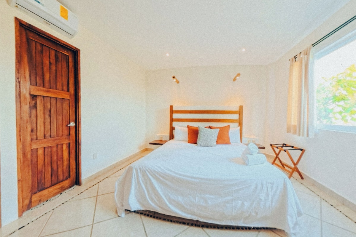 Een slaapkamer met een grote houten deur, een bed met witte lakens, een nachtkastje, een klein tafeltje en een raam met gordijnen.