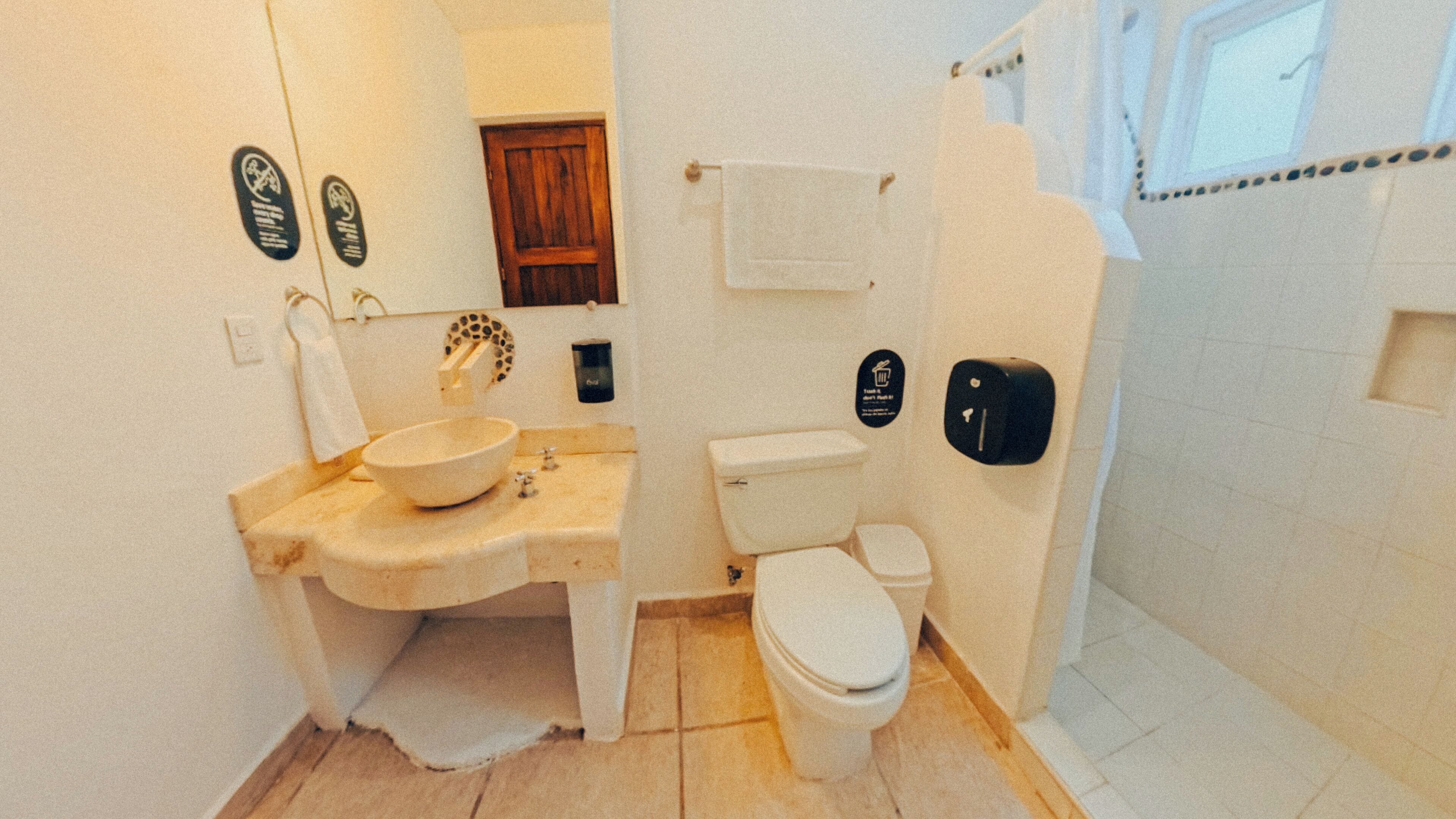 Un baño pequeño con lavamanos, inodoro y ducha. Hay carteles en las paredes y una toalla colgada en la puerta.