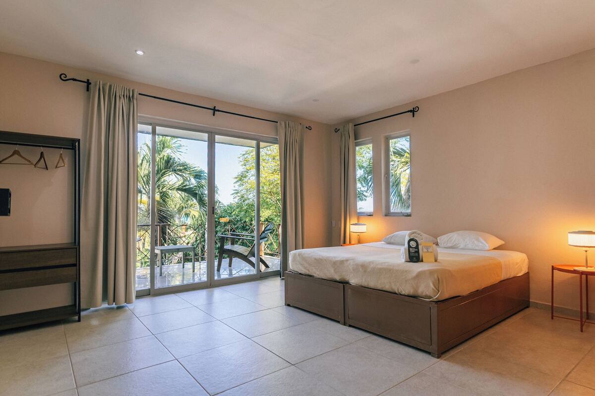 Een hotelkamer met een bed, een nachtkastje, een stoel, een raam met gordijnen en uitzicht op het buitengebied.