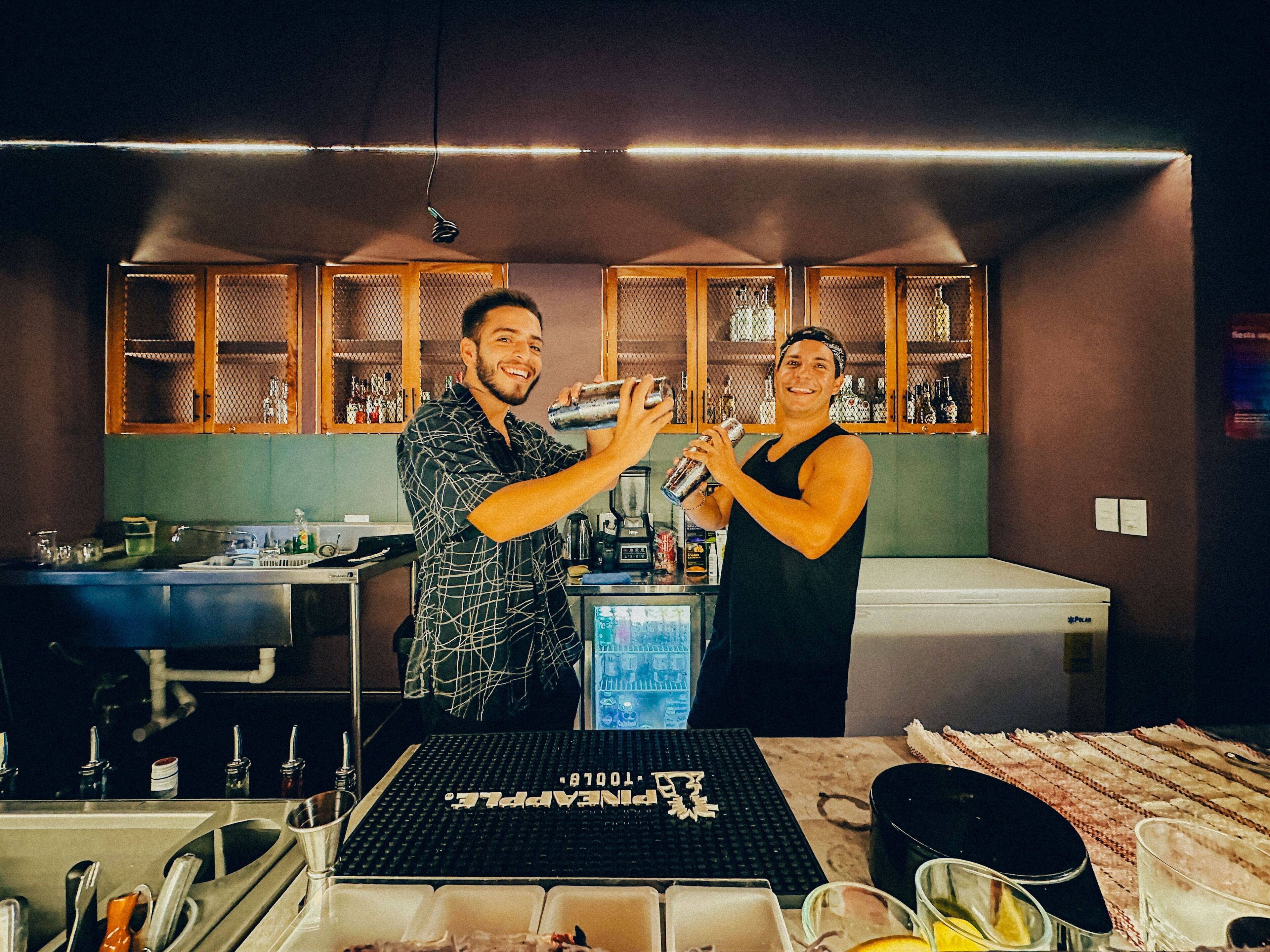 Una zona de bar con mostrador, pileta y refrigerador. Hay botellas y vasos en las estanterías detrás de la barra.