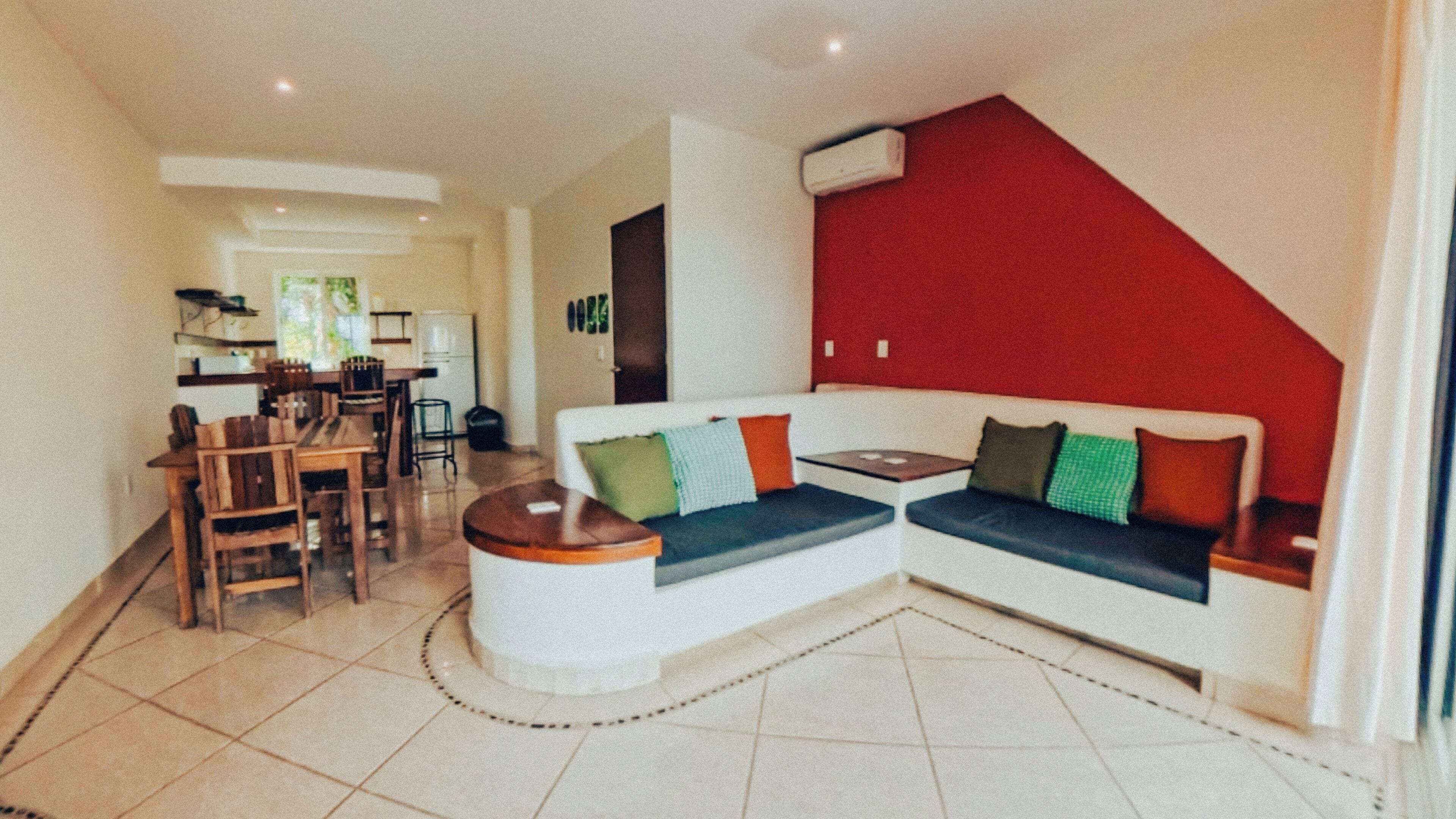 A modern living room with a white sofa, a dining area with wooden furniture, and a red accent wall.