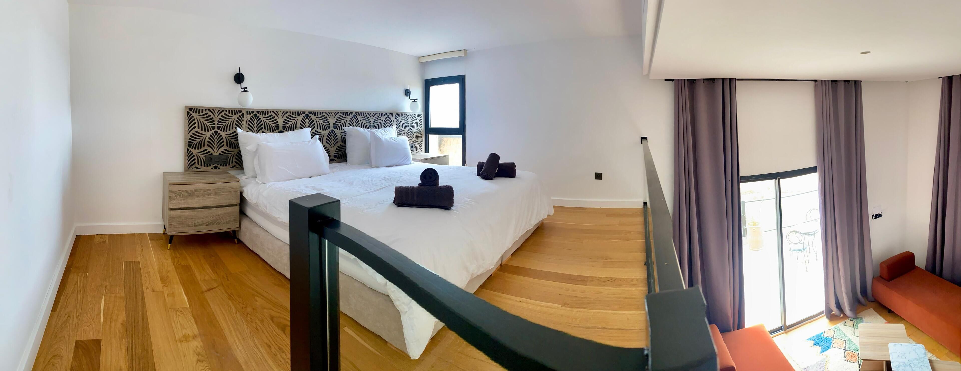 Una habitación de hotel moderna con una cama grande, suelo de madera, una mesita de noche, un escritorio y vistas al exterior.