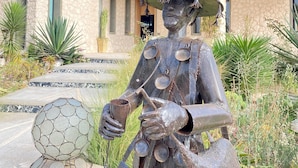 Una statua raffigura una figura che regge un annaffiatoio e una sfera di metallo, posizionata di fronte a un edificio in pietra con balconi e vasi di piante.