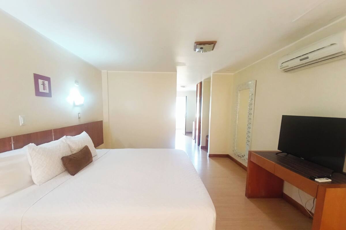 Ein Hotelzimmer mit einem großen Bett, einem Fernseher auf einem Holzständer und einer Klimaanlage an der Wand.