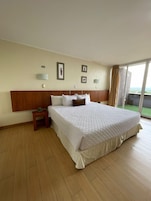 Uma cama grande com cabeceira de madeira, uma mesinha de cabeceira com uma planta e vista para o exterior por uma janela ampla.