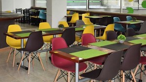 Een cafetaria met kleurrijke stoelen en tafels, een centraal bedieningsschuif en een uitzicht op buiten door grote ramen.