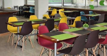 Uma cafeteria com cadeiras e mesas coloridas, um balcão central e vista para o exterior por grandes janelas.