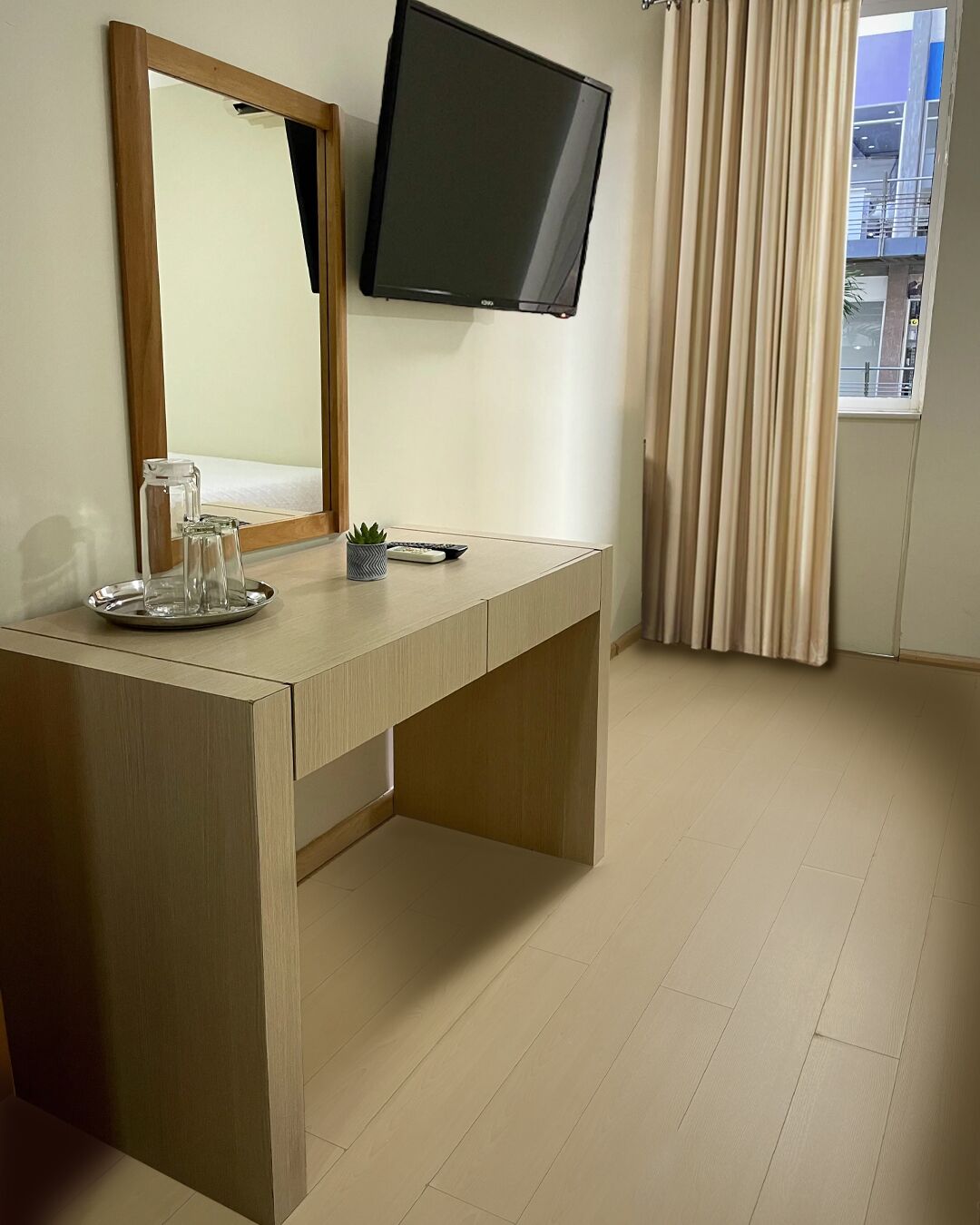 Een hotelkamer met een houten bureau, een flatscreen televisie, een spiegel en een raam met gordijnen.