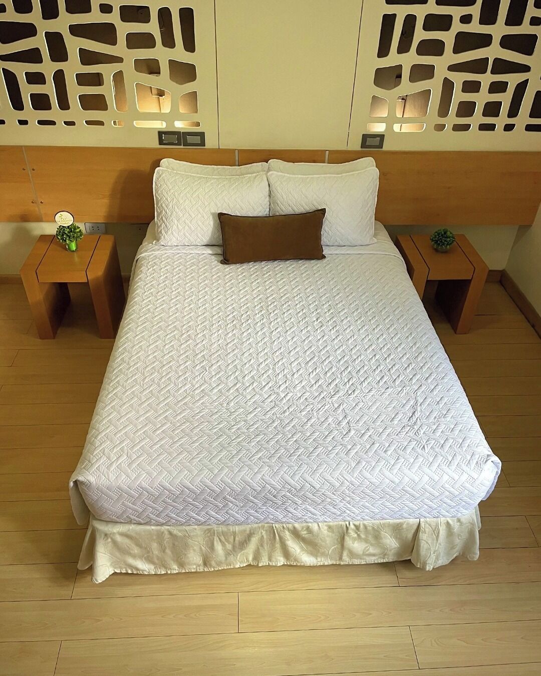 A neatly made bed with a white quilt and a brown pillow, flanked by two nightstands with small green plants.