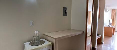 A hotel room with a compact kitchenette, a wooden floor, a light-colored wall, and a ceiling light.