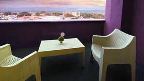 A balcony with two wicker chairs and a small table, overlooking a cityscape during sunset.