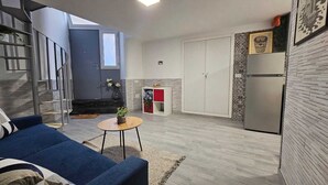 Un espace de vie compact comprenant un canapé bleu, une petite table ronde avec une plante, un coin cuisine avec un réfrigérateur et un escalier menant à un niveau supérieur.