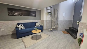 A living room with a blue sofa, a round coffee table, a spiral staircase, and a cityscape painting on the wall. - In the beating heart of the city-historical center (Napoli)
