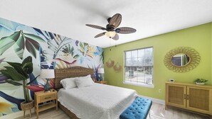 Een slaapkamer met een bed, een plafondventilator, een raam met jaloezieën, een wandspiegel en een decoratief behang.