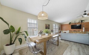 Een ruime woonkamer met een houten eettafel, transparante stoelen, een grijze bank en een aan de muur bevestigde televisie.
