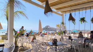 Un café situé en bord de mer, avec des chaises et des tables en osier blanc, offrant une vue sur la plage et un paysage urbain historique.