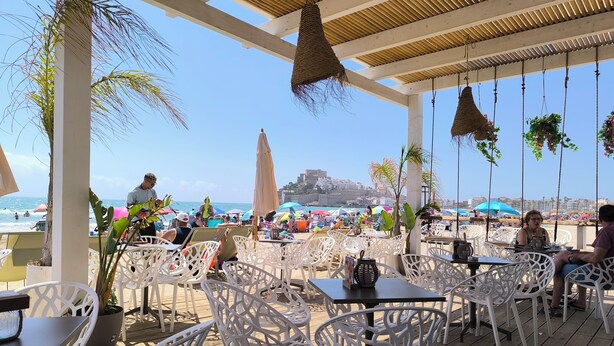 Een café aan het strand met witte rieten stoelen en tafels, met uitzicht op het strand en een historische stad.