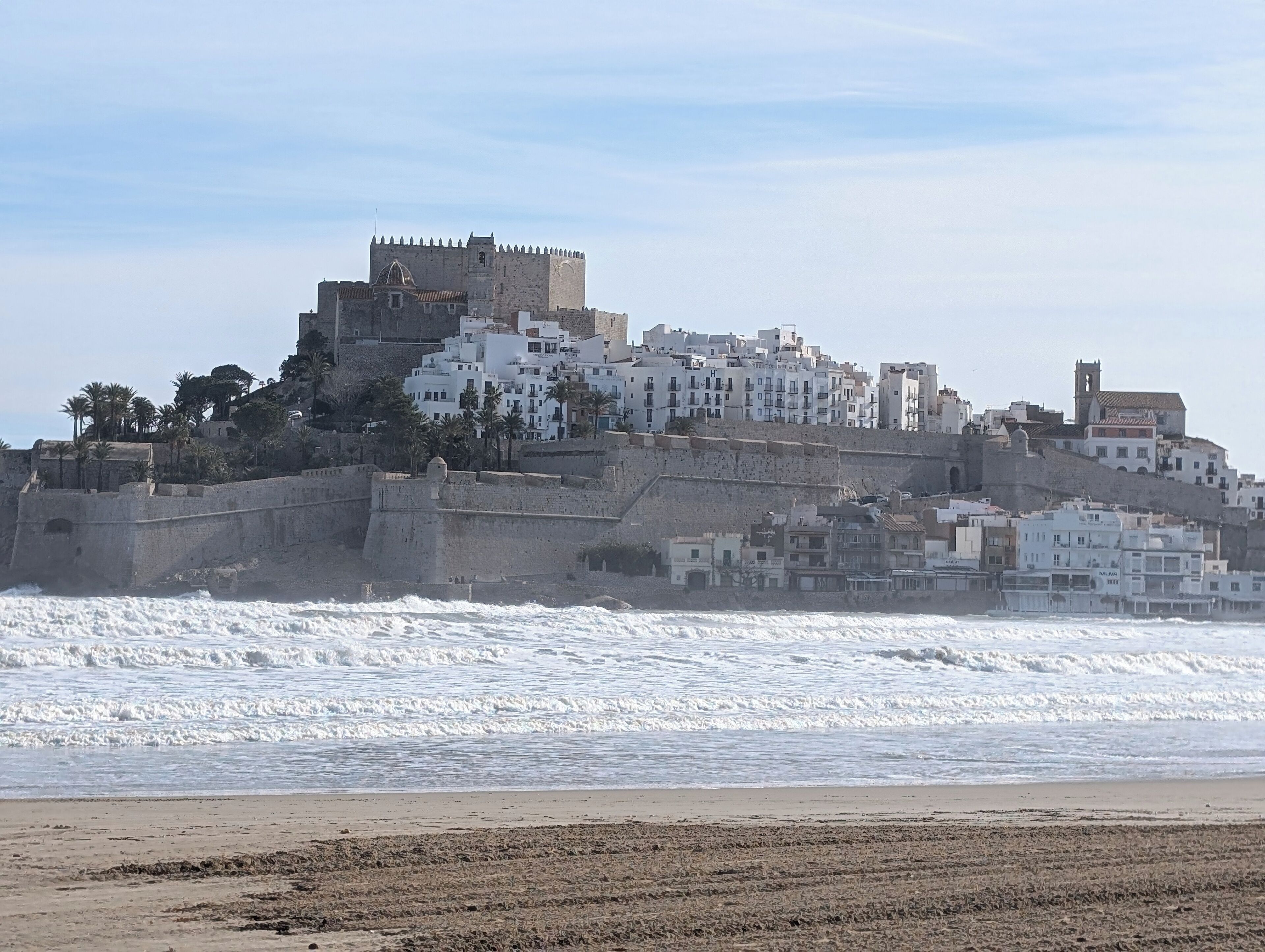 Un pueblo costero con un castillo fortificado en una colina, rodeado de edificios blancos, y olas rompiendo en la orilla.