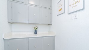 Un baño con armarios grises claros, encimera blanca y un pequeño jarrón con flores. Hay dos letreros enmarcados en la pared.