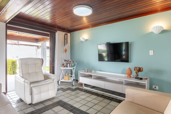 A living room with a white leather sofa, a TV mounted on the wall, and a white console table with decorative items. - Casa Luxo 5Qts 2Suítes Tamandaré SH132D (Tamandaré)