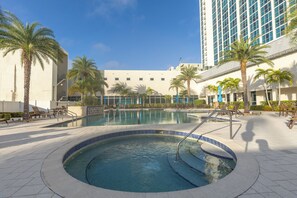 A swimming pool with a hot tub, surrounded by palm trees and lounge chairs, with a building in the background.