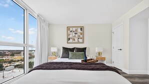 Habitación de hotel con una cama grande, dos mesitas de noche con lámparas, vista a la ciudad y una obra de arte enmarcada en la pared.
