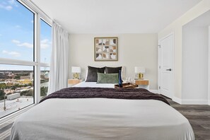 Habitación de hotel con una cama grande, dos mesitas de noche con lámparas, vista a la ciudad y una obra de arte enmarcada en la pared.