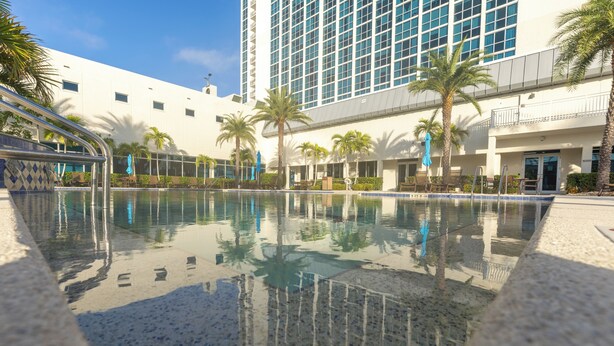 Una piscina con agua cristalina, rodeada de palmeras y un edificio moderno con grandes ventanales.