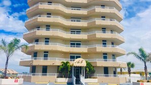 A multi-story hotel with a central entrance, balconies on each floor, and palm trees in the foreground. - Relax in Style: 2BR Ocean View Condo at Atlantic Terrace (Daytona Beach)