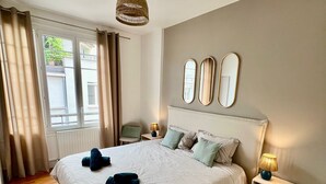 A bedroom with a large bed, a bedside table with a lamp, a chair, and a window with curtains.