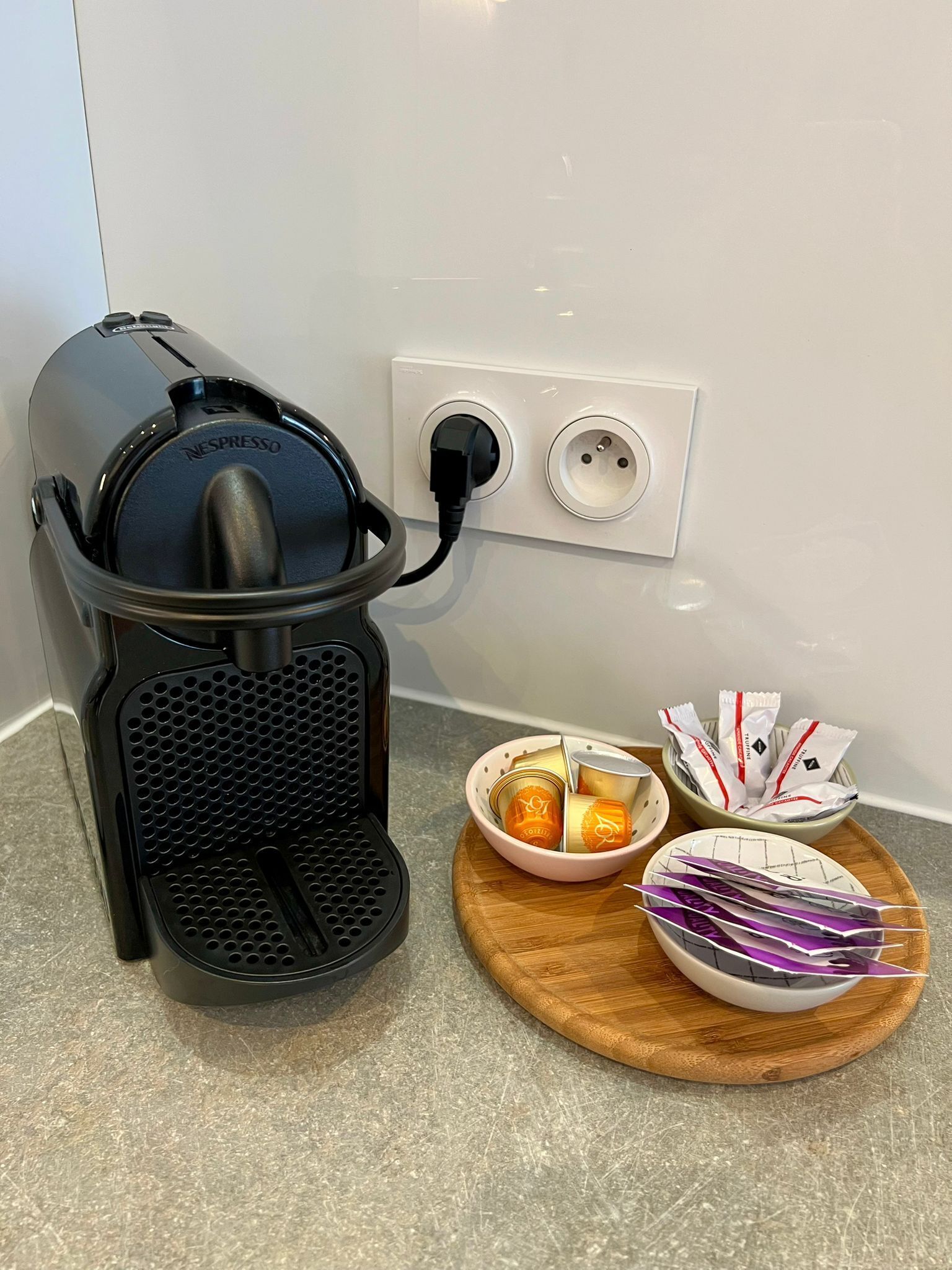 A Nespresso coffee machine on a countertop with a power outlet, a bowl of coffee capsules, and a tray with coffee pods.