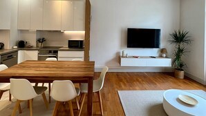 A modern living room with a wooden dining table, white chairs, a flat-screen TV, a potted plant, and a round white coffee table.