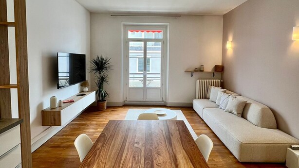 A modern living room with a wooden dining table, white sofas, and a TV mounted on the wall.