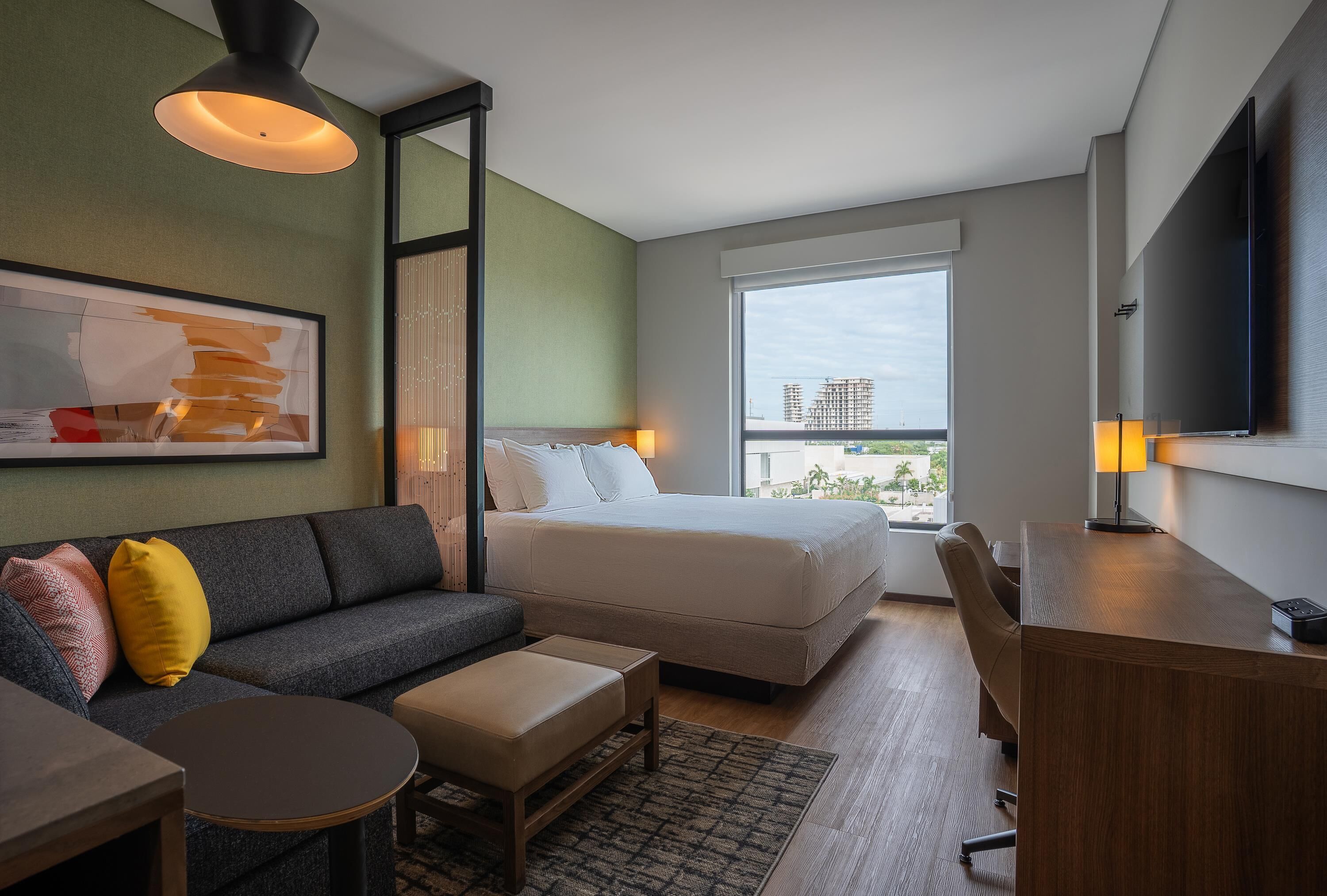 A modern hotel room with a bed, sofa, desk, and a large window offering a city view.