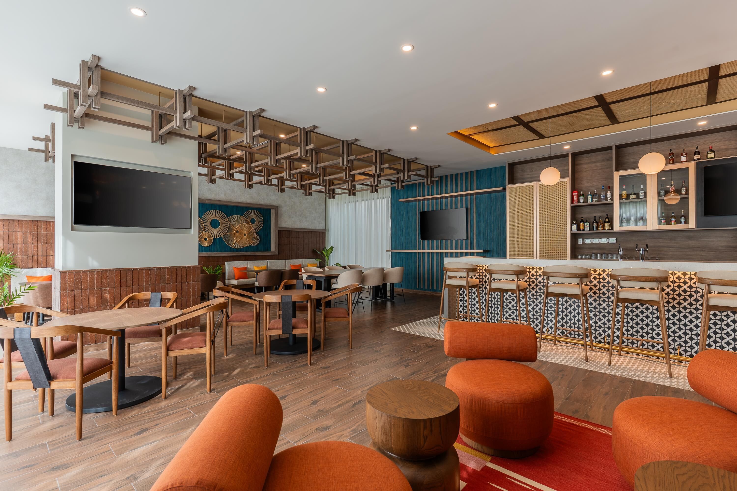 A modern restaurant with wooden tables and orange seating, a bar area with shelves of bottles, and a large TV mounted on the wall.