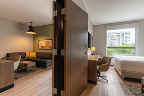 A modern hotel room with a wooden desk, a sofa, a bed, and a large window offering a city view.