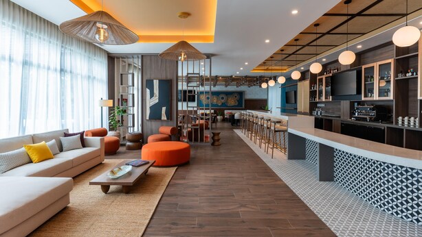 A modern hotel lobby with a sofa, ottoman, and coffee table. There is a bar area with stools and a television. - Hyatt Place Merida/Via Montejo (Mérida)