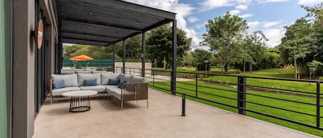 Um pátio externo com área de estar coberta, vista para um parque e gramado verde.