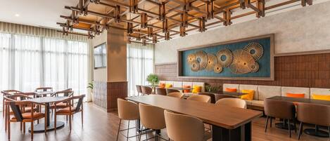 A modern dining area with a long wooden table and chairs, a high ceiling with exposed beams, and a large abstract wall art piece.
