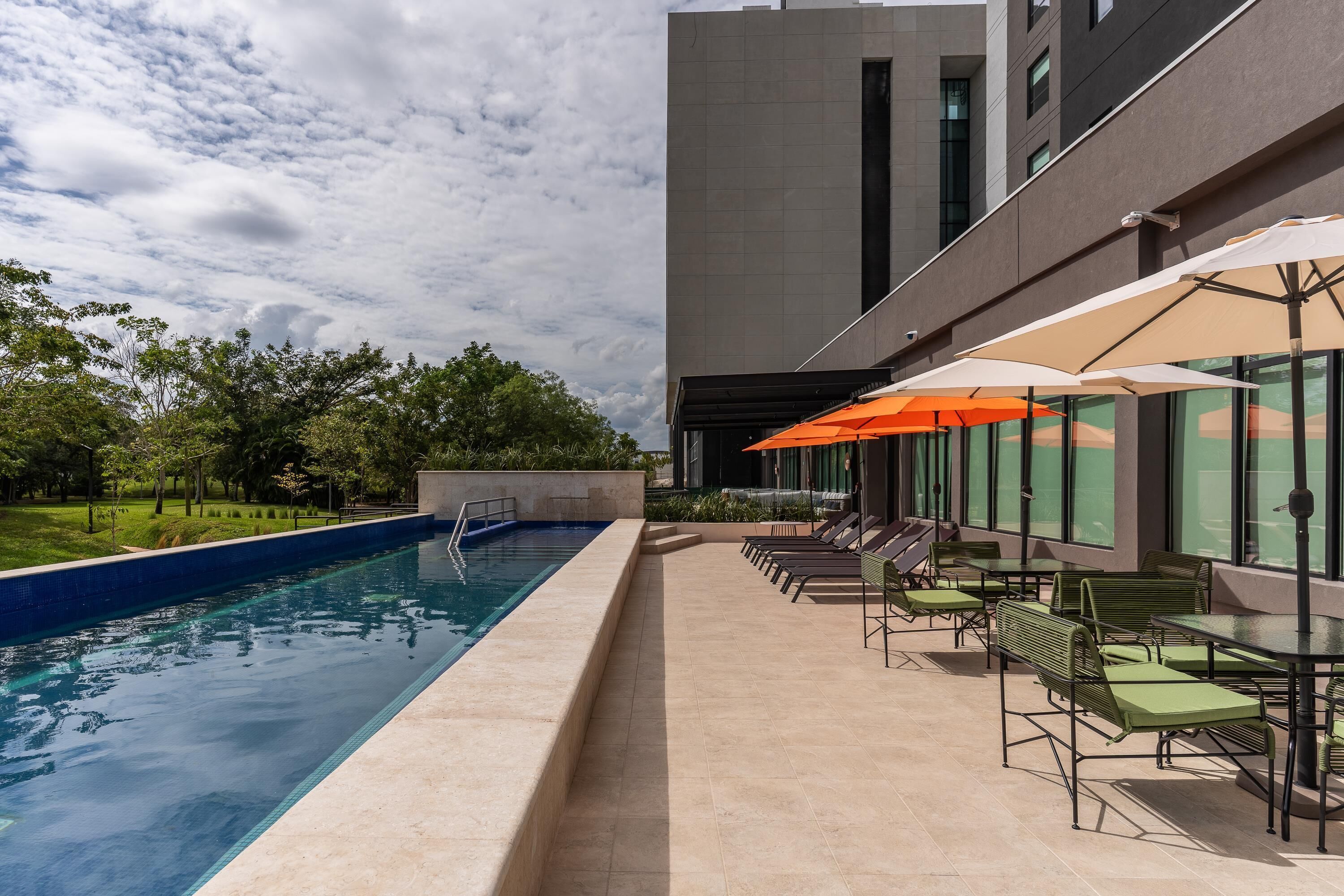 Una piscina con sillas de descanso y sombrillas, rodeada por un edificio moderno con grandes ventanales.