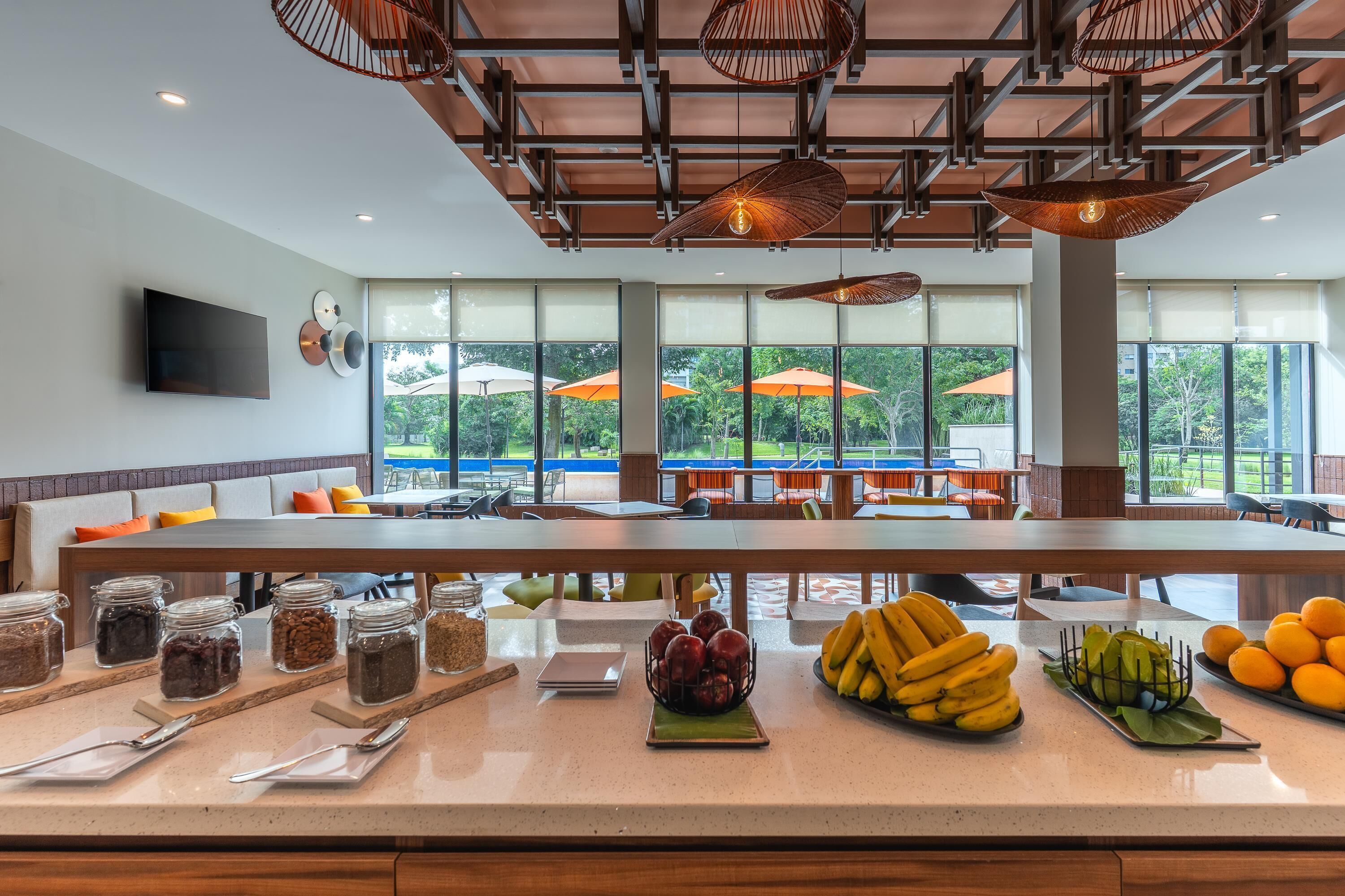 A breakfast bar with fruit and condiments, large windows with outdoor umbrellas, and a modern ceiling design.