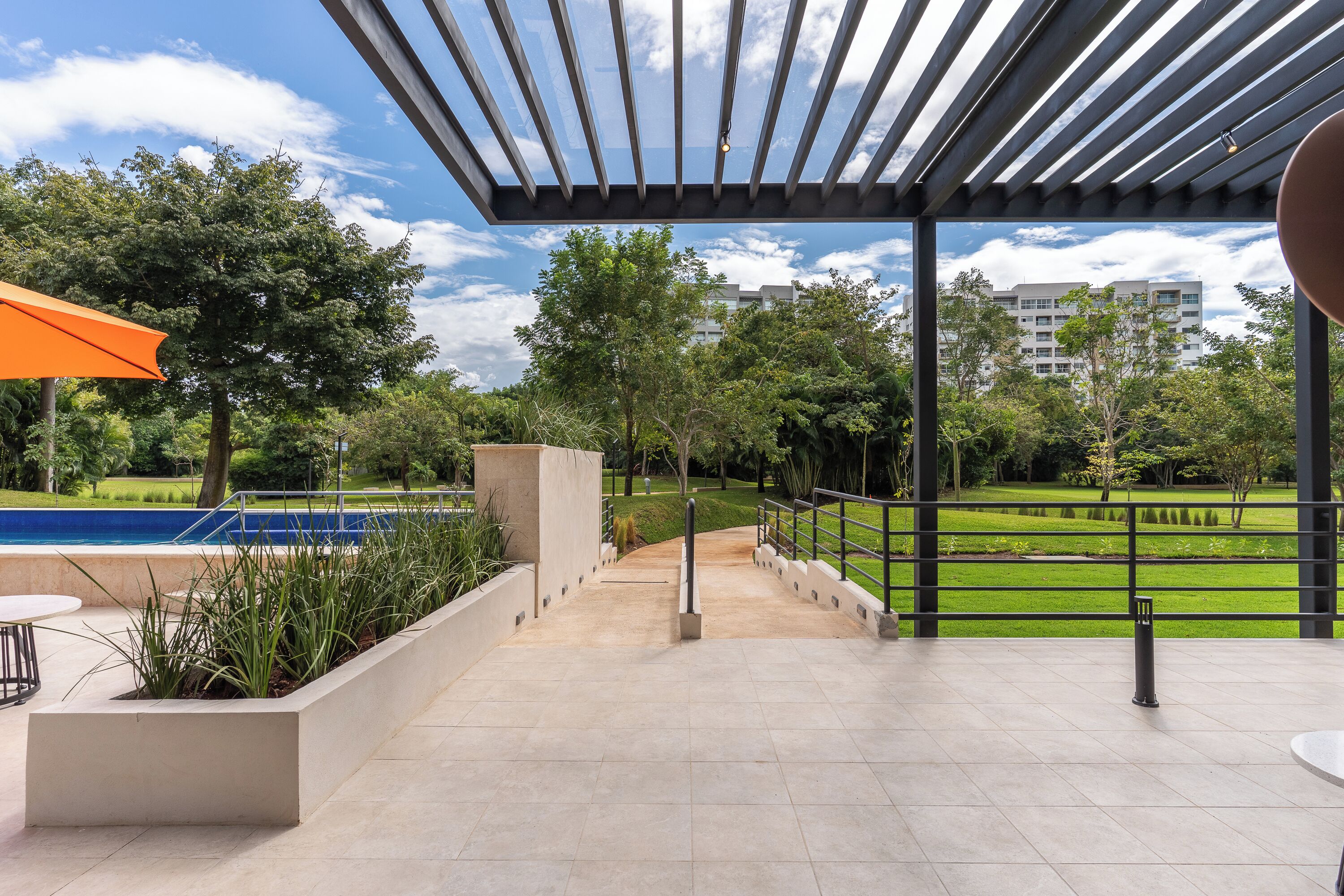 Un patio exterior con piscina, un pérgola y vistas a un parque y edificios al fondo.