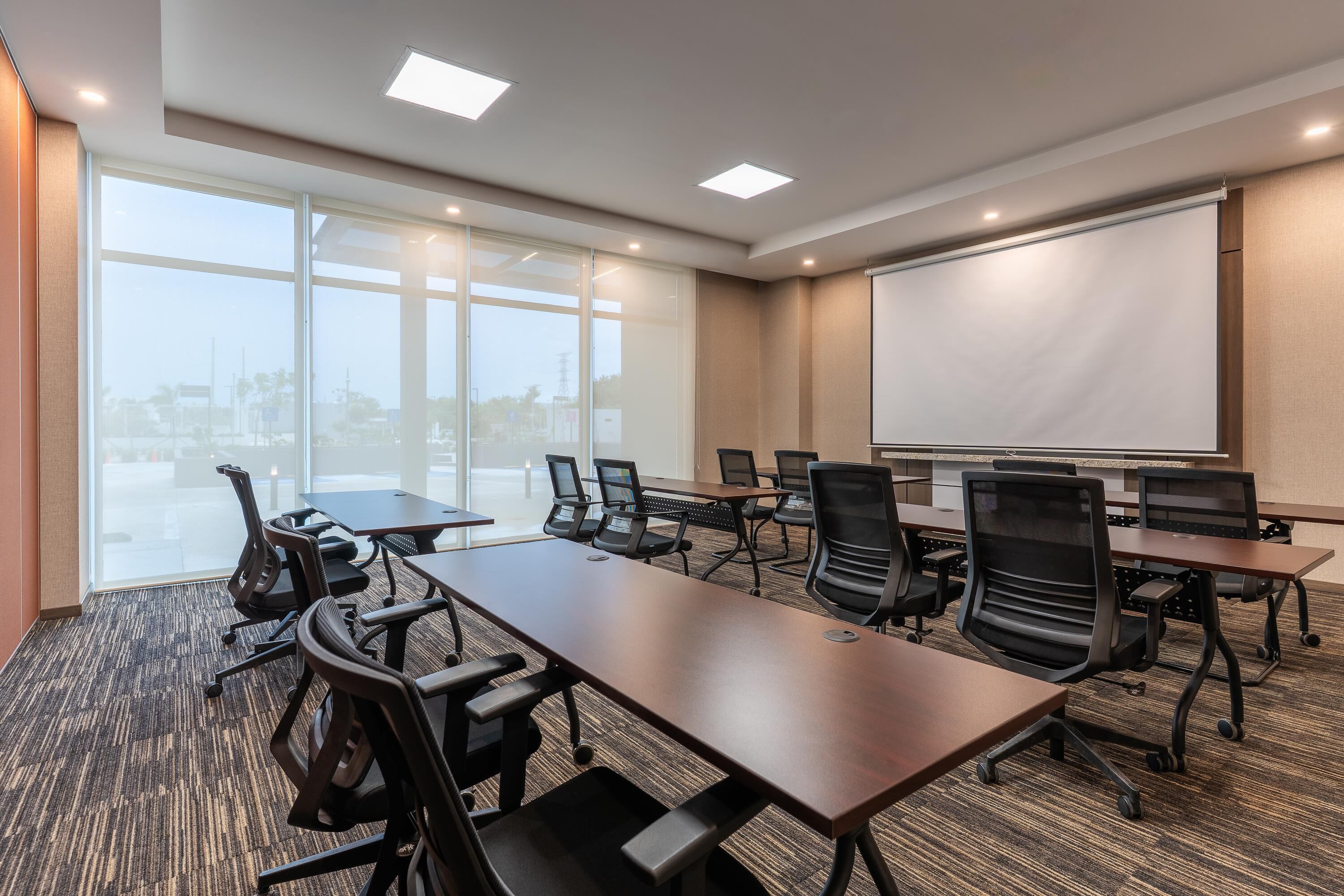 A conference room with a long table, multiple chairs, a projector screen, and large windows.