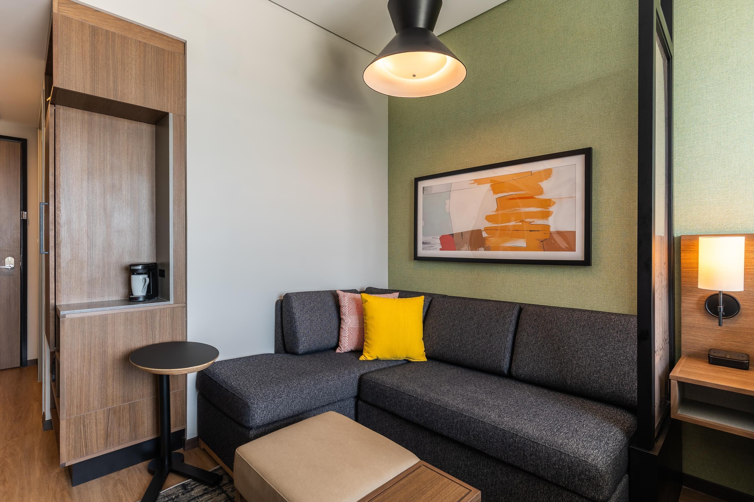 A modern hotel room with a grey sofa, a small round table, a wooden cabinet, and a framed abstract painting on the wall.