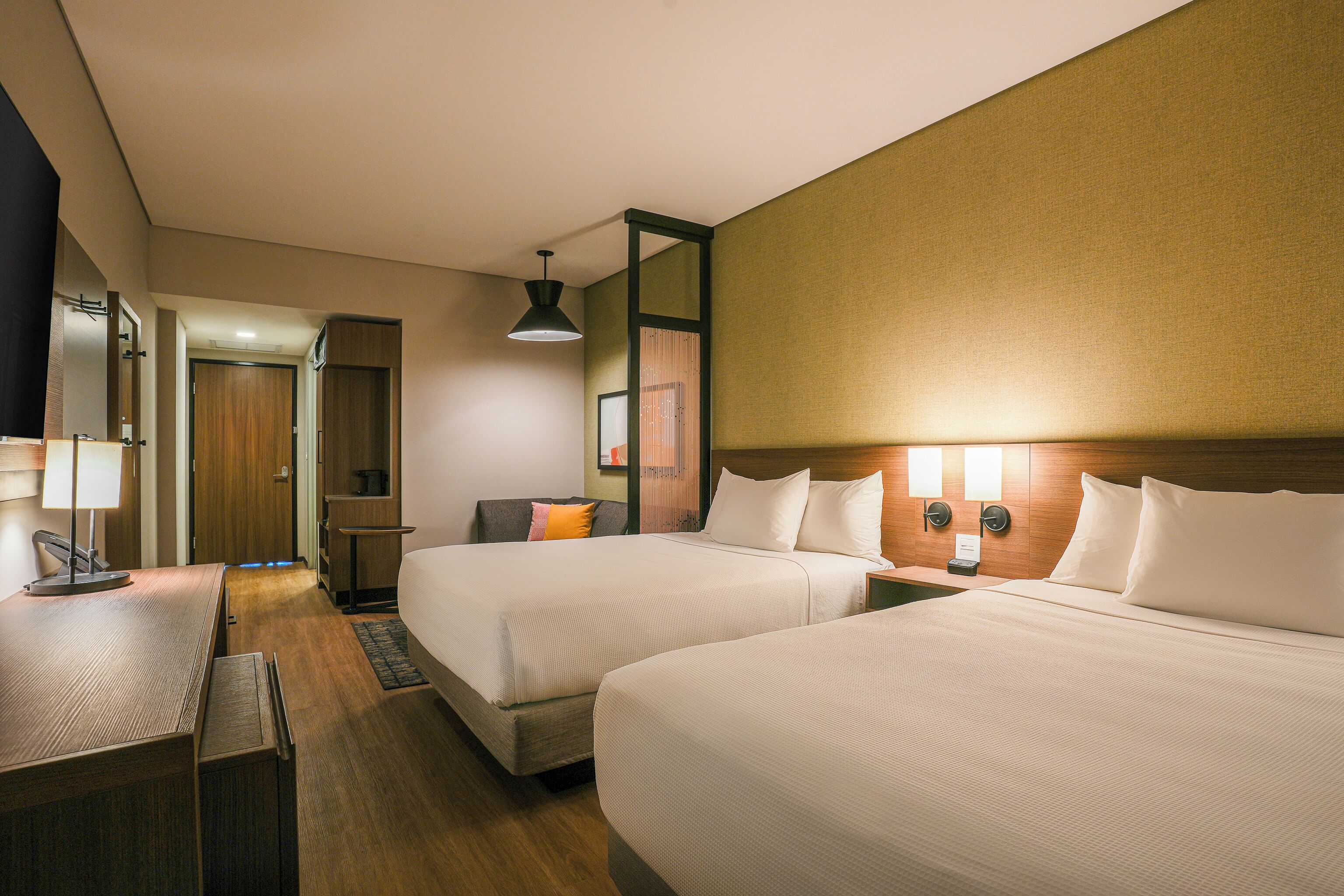 A hotel room with two beds, a desk, a lamp, and a mirror.