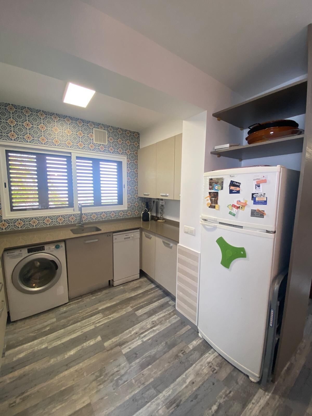 A compact kitchen with a washing machine, a refrigerator, and a window with blinds.