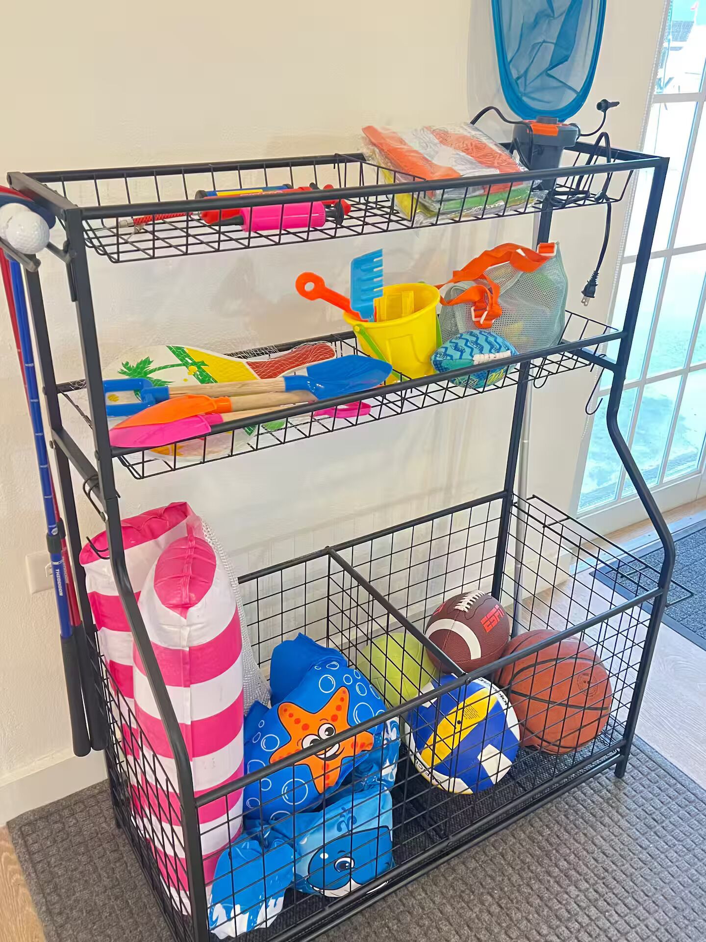 Una estantería metálica negra con varios objetos, entre ellos palas de colores, un cubo amarillo y balones deportivos.