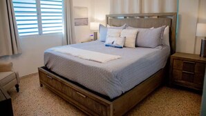 Un lit bien fait, avec un cadre en bois et une couette blanche, une table de chevet avec une lampe, et une fenêtre avec des stores.