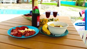 Une table en bois sur laquelle on trouve une assiette de charcuteries, un bol de bananes coupées en tranches, une bouteille de vin et deux verres de vin rouge.